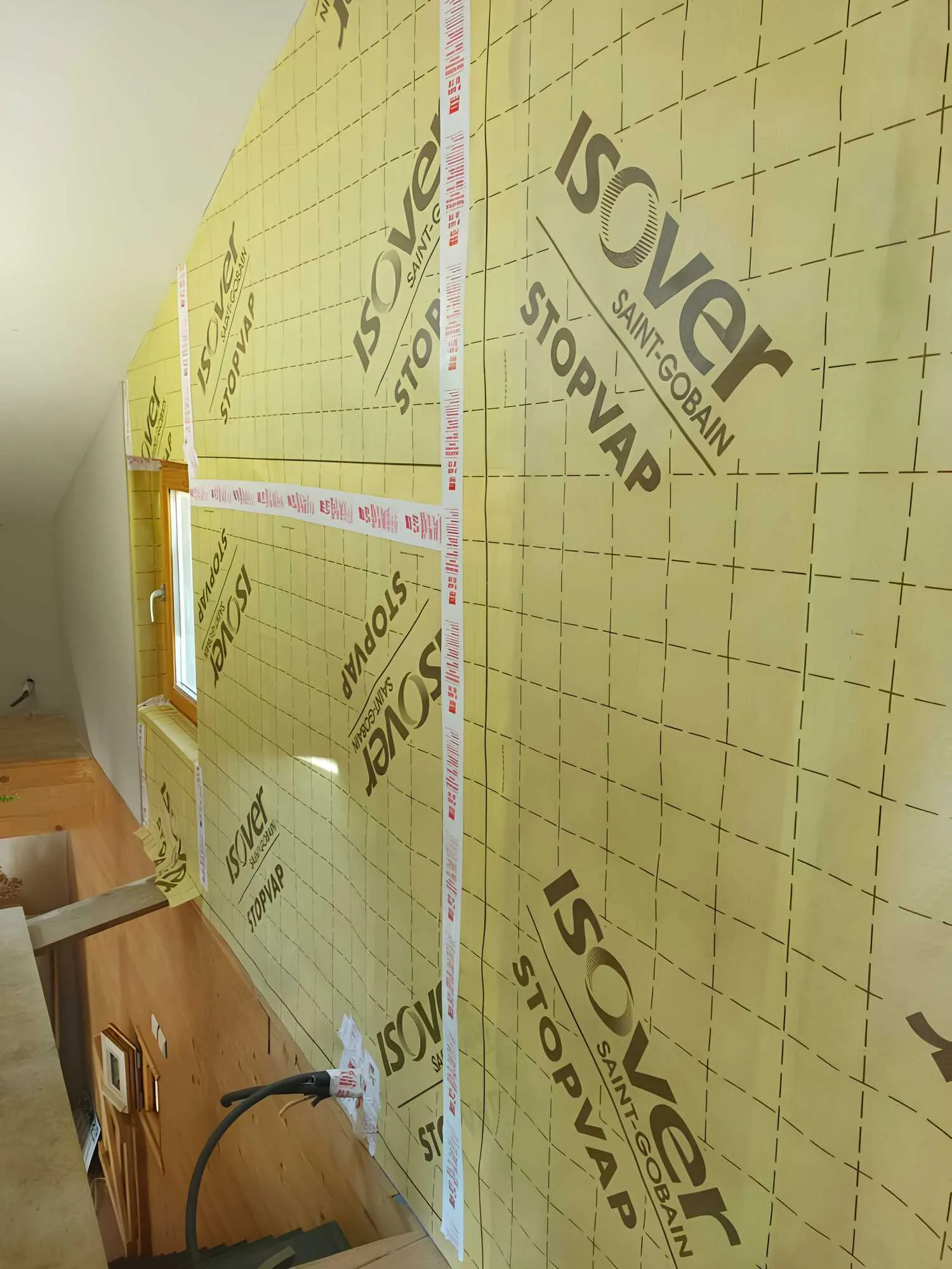 Paroi sous pente couverte d’une membrane technique jaune autour d’une fenêtre en bois.
