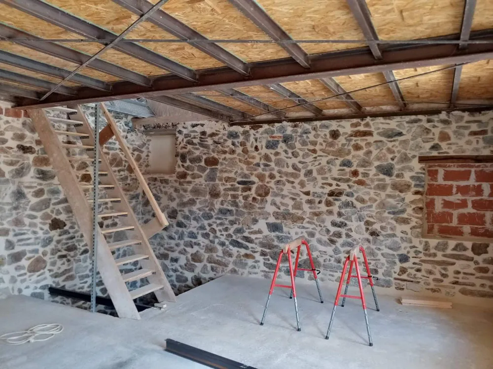 Chantier de rénovation intérieure avant pose de placo, avec structure métallique de plafond et murs en pierre apparente.