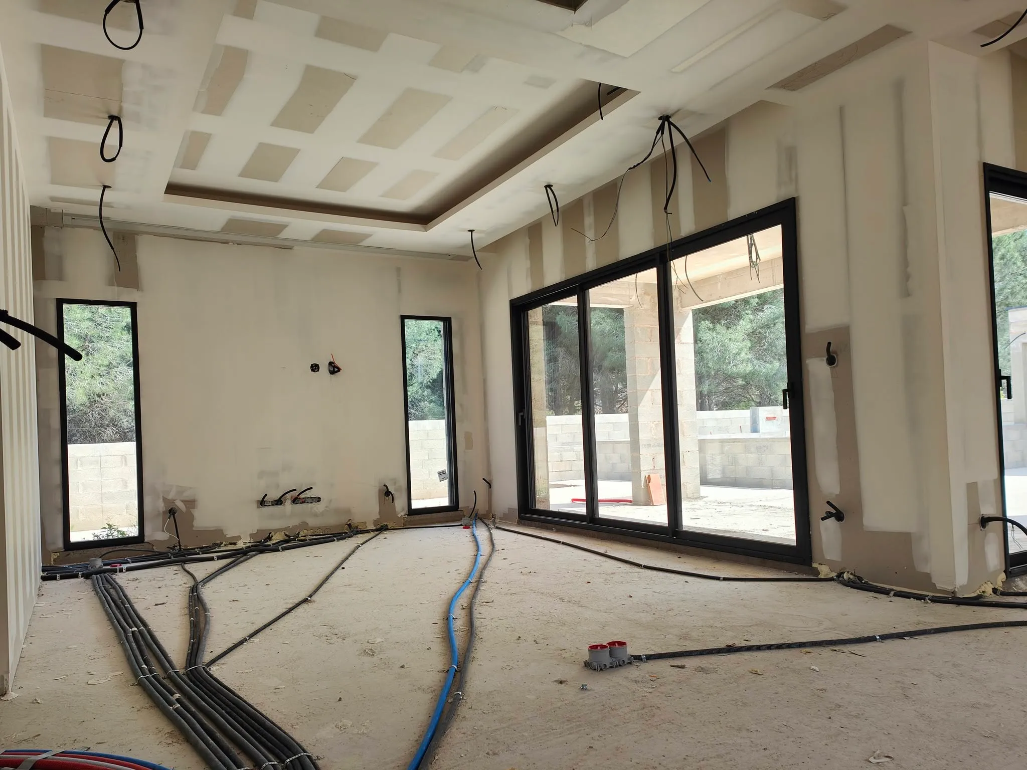 Séjour d’une villa neuve en chantier avec plafond en plaques de plâtre en cours de jointoiement, baies vitrées noires et gaines électriques au sol.