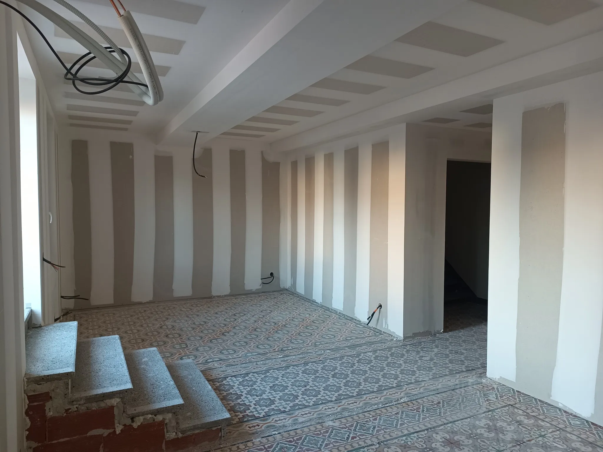 Pièce en cours de plâtrerie avec plafond en plaques, cloisons jointoyées, carreaux à motifs au sol et marches en pierre
